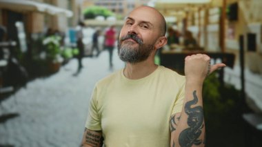 Şehir sokaklarında sakallı, dövmeli sıradan kıyafetler giyen bir adam açık şehir ortamında kendinden emin ve odaklanmış görünüyor..