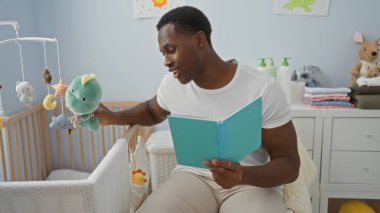Yatak odasında yakışıklı bir adam beşikteki bebeğe pelüş oyuncaklarla kitap okuyor ve sıcak bir atmosfer yaratıyor.