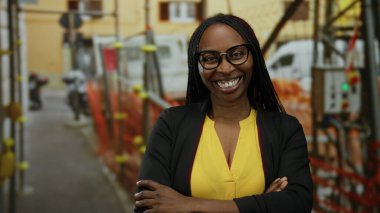 Açık hava inşaat alanında sarı tişörtlü, kıvırcık saçlı, gülümseyen kadın, yapı iskelesi ve ekipmanlar arasında mutluluk ve güven gösterisi yapıyor..