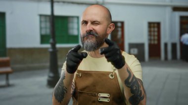 Sakallı ve dövmeli bir adam önlük takıyor ve eldiven takıyor. Arka planda bulanık binalar olan şehir caddesindeki kameraya bakıyor..