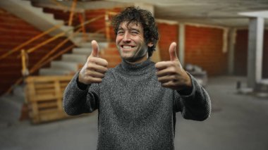 İnşaat alanında gülümseyen genç bir adam sıradan bir kazakla başparmağını kaldırıyor.