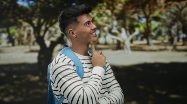 Güneşli bir günde açık hava parkında sırt çantasıyla gülümseyen genç bir adam..