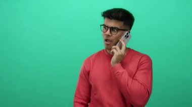 Gözlüklü genç adam yeşil arka planda telefonda konuşuyor, kırmızı kazak giyiyor, sürpriz ve jest yapıyor, iletişimi ve duyguyu vurguluyor..