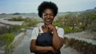 Önlüklü, kendine güvenen bir kadın, açık bir gökyüzünün altında, doğal yeşillik ve geniş açık alanlarla çevrili, sıcaklığı ve dostluğu yayarak bir park ortamında duruyor..