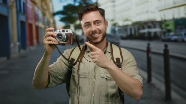 Sakallı ve sırt çantalı genç adam canlı bir şehir caddesinde kameraya gülümseyip işaret ederek turist ve fotoğrafçı olarak hevesini sergiliyor..