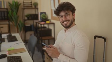 Modern ofis ortamında akıllı telefon kullanan sakallı genç adam dijital teknoloji, bitkiler ve ofis mobilyalarıyla çevrili, profesyonel bir iş ortamı sunuyor..