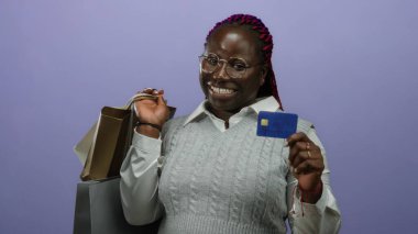Alışveriş torbaları ve kredi kartı tutan bir kadın stüdyo ortamında izole edilmiş mor bir arkaplanda neşe ve tüketim göstergesi olarak gülümsüyor..