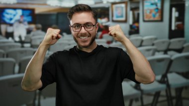 Sakallı ve gözlüklü genç adam modern sanat galerisindeki yumruklarıyla güvenle gülümsüyor. Etrafı çeşitli sanat eserleri ve boş sandalyelerle çevrili..