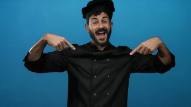 Şef coşkuyla kendini gösteriyor, parlak mavi bir arka plana karşı siyah bir üniforma giyiyor, aşçılık uzmanlığına karşı gurur ve güven gösteriyor..