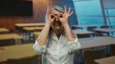 Gri saçlı, gözlüklü, sınıfın içinde jest yapan, neşe ve yaratıcılığını ifade eden yaşlı bir kadın..