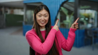 Pembe kazaklı kadın gülümsüyor ve modern mavi tententenin altında heyecanlı bir şekilde bir şehir sokağını işaret ediyor, genç ve neşeli tavırlarını vurguluyor..