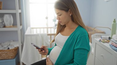 Hamile kadın rahat bir yatak odasında oturmuş karnını sarmalayıp, akıllı telefonuna bakıyor. Etrafı yumuşak iç dekorasyon ve bebek eşyalarıyla çevrili..