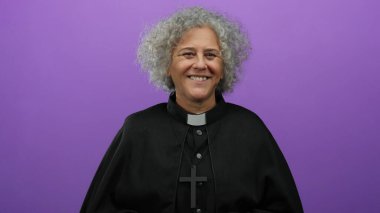 Siyah papaz kıyafeti giymiş gri saçlı yaşlı kadın sırıtışına işaret ederken gülümsüyor, canlı mor bir duvara yaslanıyor, neşeli ve cana yakın bir tavır sergiliyor..