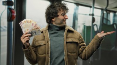 Hispanik bir adam otobüsün içinde Türk lira banknotları sunuyor, hareketli bir jest yapıyor, şehir içi ulaşım sistemindeki canlı ekonomik değişimi vurguluyor..