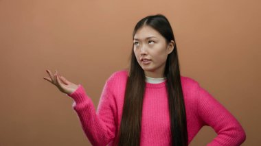 Kahverengi duvar arkasına karşı duygularını ifade eden pembe kazaklı, soyutlanmış Asyalı kadın pozu veren bir kadın..