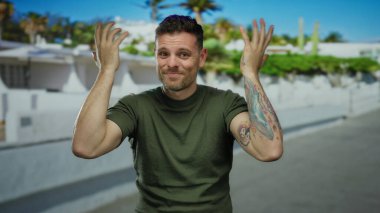 Dövmeleri olan İspanyol bir adam sokak ortamının dışında duruyor, elleri başının üstünde endişeli görünüyor, yeşil bir gömlek giyiyor ve duygularını ifade ediyor..