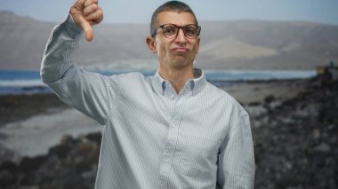 Sahilde, güneşli bir havada, açık deniz arka planına karşı, baş parmağıyla jest yapan, gözlük takan ve sıradan gömlekler giyen bir adam..