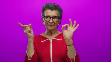 Gri saçlı, gözlüklü, kırmızı kazaklı yaşlı bir kadın canlı pembe arka plana karşı iyi bir jest yapıyor. Pozitiflik ve kendine güveni ima ediyor..