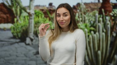 Beyaz jestli kadın, arka planda kaktüsüyle dışarıda küçük boy jest yapıyor. Park ortamında neşeli bir ifade sergiliyor..