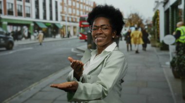 Genç bir kadın, kırmızı otobüsler ve müşterilerle dolu kalabalık bir şehir caddesinde elini kameraya uzatıyor, gülümsüyor, şehir yaşamının enerji ve çeşitliliğini yakalıyor..