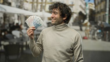 Güneşli bir şehir caddesinde, arka planda kalabalık bir açık hava restoranı ve kahve dükkanı terası olan, Polonyalı zloti banknotlarını tutarken gülümseyen İspanyol adam, şehir hayatının özünü yakalıyor..