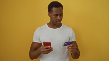 Akıllı telefon ve kredi kartı tutan genç adam izole edilmiş sarı arka plana karşı duruyor. Odaklanma ve konsantrasyon gösteriyor.