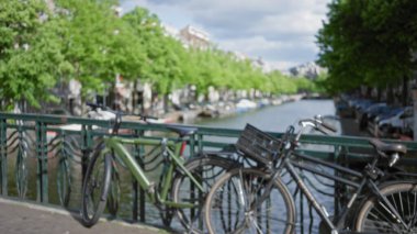 Bulanık bisikletler Amsterdam 'daki bir kanal köprüsünde duruyor. Etrafı yemyeşil yemyeşil, Amsterdam' ın manzaralı su yolları ve canlı şehir atmosferinin özünü yakalıyor..