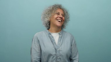 Gri saçlı bir kadın, izole edilmiş mavi bir duvara sevinçle gülüyor. Rahat bir poz vererek mutluluk ve kendine güven yayıyor. Orta yaşlı zarafetini sergiliyor..