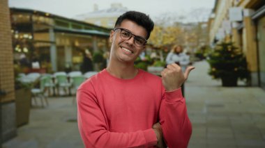 Şehir sokaklarında gülümseyen gözlüklü genç bir adam açık hava kafesinin bulanık arka planına sahip ve gündüzleri yürüyen insanlar şehir yaşam tarzını ve günlük modasını gözler önüne seriyor..