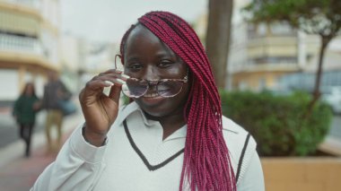 Güneşli bir günde arka planda binalar ve yeşillikler olan bardakları kaldırırken, kadın sokakta gülümsüyor..