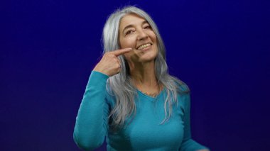 Gri saçlı yaşlı bir kadın, izole edilmiş mavi bir arka plana sıcak bir şekilde gülümsüyor, rahatlatıcı hareketlerle neşeli yüzüne işaret ediyor, kendine güvenen, huzurlu bir anı yakalıyor..