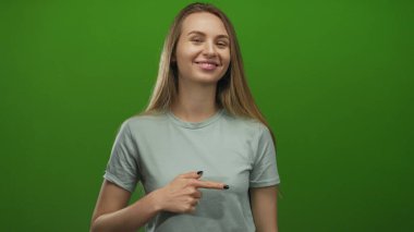 Gülümseyen ve işaret eden bir kadın canlı yeşil izole bir arka plan karşısında, pozitiflik ve güven yayarak, günlük tarzını ve anlamlı jestini vurguluyor..