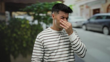 Gün boyunca şehir caddesinde dikilirken burnunu kapatan çizgili tişörtlü genç adam..