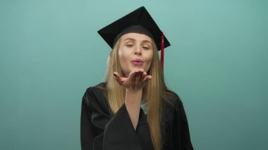 Şapkalı ve cüppeli mezun bir kadın, izole edilmiş yeşil bir duvar arkasına öpücük yolluyor, neşe ve sevinç yakalıyor..