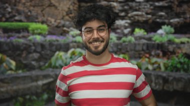 Gözlüklü ve sakallı genç adam arka planda yemyeşil bir parkta çizgili bir gömlek içinde gülümsüyor.