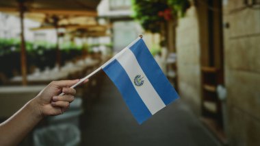 El ele tutuşan Salvador bayrağı, bulanık kafenin arka planında, kasabanın huzurlu bir sokak atmosferini yakalıyor..