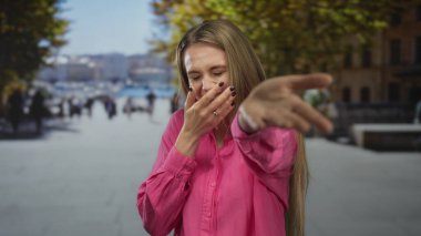 Kadın hareketli bir şehir caddesinde gülüyor ve kamerayı işaret ediyor. Canlı duygular ve şehir enerjisi sergiliyor. Arka planda yoldan geçenler ve yol boyunca ağaçlar var..