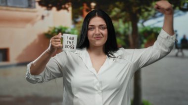 Patron bardağını dışarıda tutan kendine güvenen bir kadın gün ışığında kentsel çevrede güç gösterisi yapıyor. Arka planda ağaçlar pozitiflik ve kendine güven gösteriyor.