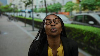 Kıvırcık saçlı ve gözlüklü gülümseyen kadın, şehir sokağında elleri açık, canlı sarı gömlekli, neşe ve pozitiflik saçan bir kalp yapar..