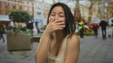 Şehir arka planında bulanık şehir arka planında, açık havada gülen bir kadın aydınlık bir günde gençlik neşesi ve günlük giysiler sergiliyor ve insanlar uzaklarda yürüyor..