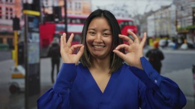 Kalabalık bir caddede arka planda kırmızı bir otobüsle mavi gömlekli ve açık havada gülümseyen Çinli bir kadın iyi bir jest yapıyor..