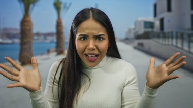 Sahilde güneşli bir gezinti yaparken, arka planda palmiye ağaçları ve modern binalar olan bir kadın, açık hava stresi hissi veriyor..