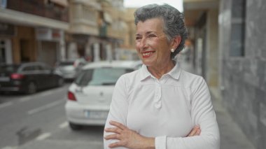Gri saçlı ve çapraz kollu bir kadın şehir sokağında arka planda arabalarla samimi bir şekilde gülümsüyor, beyaz giyinmiş, neşeli ve dostane bir açık hava sahnesini vurguluyor..