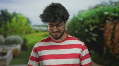 Sakallı, gözlüklü, çizgili tişörtlü, yeşil ve bitkilerle çevrili bir parkta duran genç bir adam.