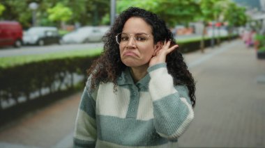 Kıvırcık saçlı ve gözlüklü İspanyol kadın sokakta dikiliyor, eli kulağında, yeşillik ve araçlarla çevrili bir şekilde dinlemek için işaret ediyor..