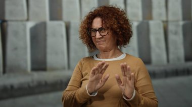 Orta yaşlı, kıvırcık kahverengi saçlı İspanyol kadın şehir sokaklarında gözlük ve kahverengi süveter takıp duygularını dışa vurarak bir jest yapıyor..
