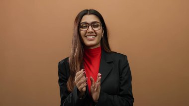 Düz kahverengi arkaplana karşı iş elbisesi içinde sevinçle alkışlayan bir kadın profesyonel bir ortamda güven ve başarı sergiliyor..