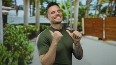 Dövmeli genç adam gülümsüyor ve yeşil arka planda ağaçlar ve yeşil bir şehir ortamında başparmak kaldırıyor, pozitiflik ve güven taşıyor..
