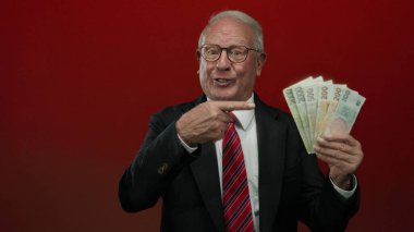 İş kıyafeti giymiş kıdemli bir adam, elinde Czech Koruna banknotları ile izole edilmiş kırmızı bir zemin üzerinde başarı ve zenginliği ima eden kendine güvenen bir ifadeyle..