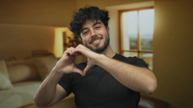 Elleri rahat otel odasında, geniş pencereli ve sıcak ışıklandırmalı kalp sembolü yapan genç adam mutluluğu ve olumlu duyguyu ifade ediyor..